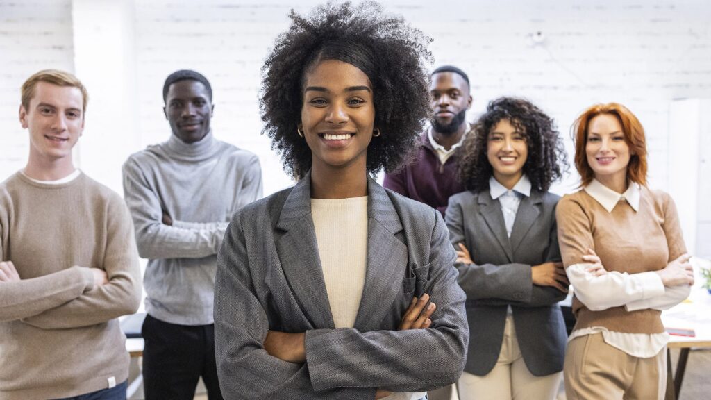 group of young multiracial business people, focus on latin american woman. Smiling young businesswoman stands confidently in front of her diverse team, arms crossed, representing leadership and teamwork.
